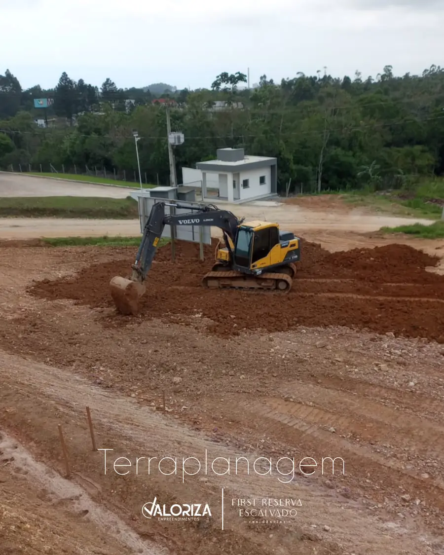 Escavadeira nivelando o terreno, com a guarita de acesso ao fundo